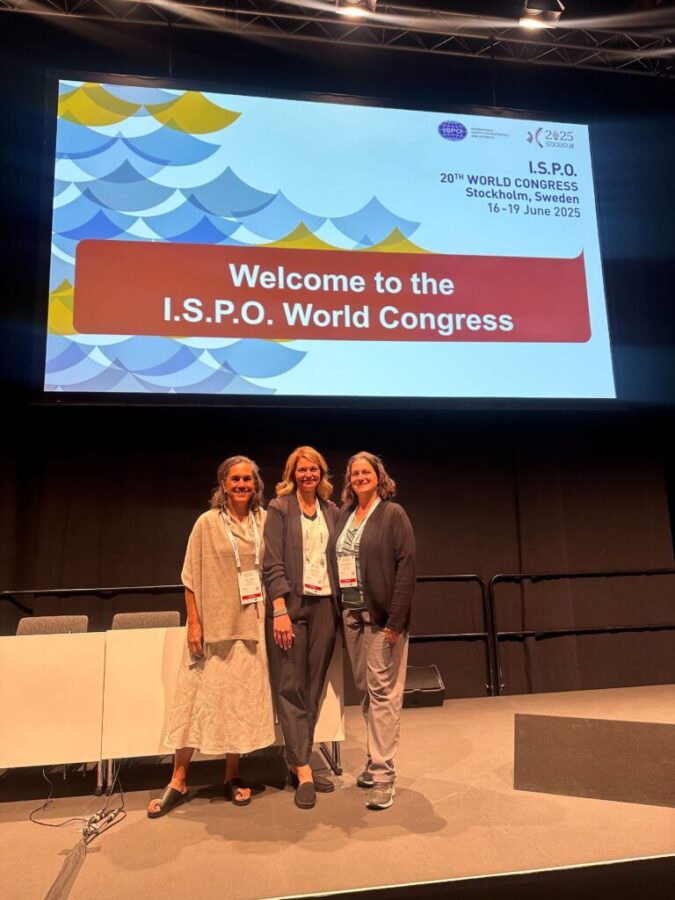 Three members of the Physiopedia team posing together on stage at the ISPO 20th World Congress in Stockholm, Sweden, standing in front of a large screen displaying the conference welcome message with colorful wave branding.